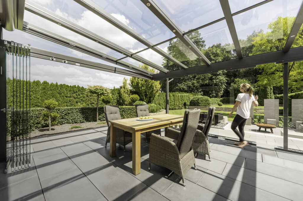 Frau schiebt die senkrechten Glas-Elemente vom Glashaus an die Seite und verwandelt es so in eine offene Terrassenüberdachung